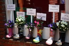 6 verschieden bunte Gummistiefel mit Blumen gefüllt stehen vor einem Fenster. In jedem Gummistiefel ist ein Schild mit "Herzlich Willkommen" auf verschiedenen Sprachen.